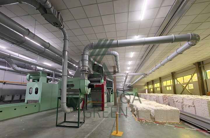 Industrial textile processing area with large ventilation pipes, machinery, and stacked cotton blocks.